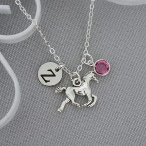 May include: A silver-toned necklace featuring a delicate chain with three charms: a circular silver disc engraved with the letter Z, a silver-toned horse pendant in a running pose, and a small, round, faceted pink crystal charm.