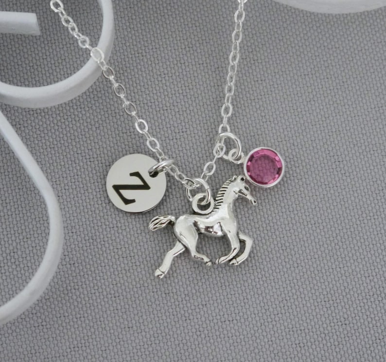 May include: A silver-toned necklace featuring a delicate chain with three charms: a circular silver disc engraved with the letter Z, a silver-toned horse pendant in a running pose, and a small, round, faceted pink crystal charm.