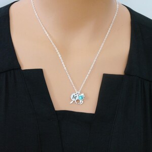 May include: A silver-toned chain necklace featuring a small circular charm engraved with the letter M, a horse-shaped charm, and a small round turquoise-colored stone charm. The necklace rests against a solid black fabric background.
