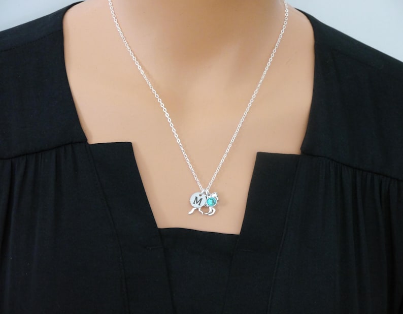 May include: A silver-toned chain necklace featuring a small circular charm engraved with the letter M, a horse-shaped charm, and a small round turquoise-colored stone charm. The necklace rests against a solid black fabric background.