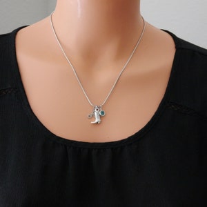 May include: A silver-toned snake chain necklace featuring three small charms: a letter A, a cowboy boot, and a round teal-coloured stone. The necklace rests against a black fabric background, showcasing a simple and delicate metallic design.