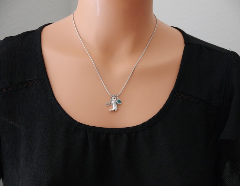 May include: A silver-toned snake chain necklace featuring three small charms: a letter A, a cowboy boot, and a round teal-coloured stone. The necklace rests against a black fabric background, showcasing a simple and delicate metallic design.