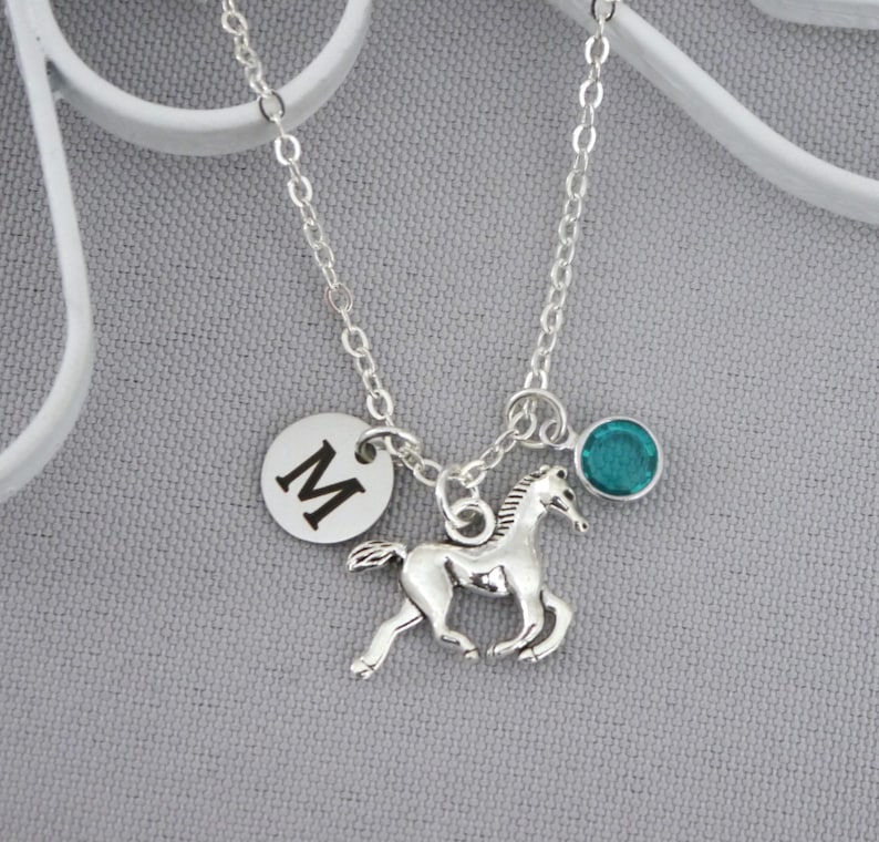 May include: A silver-toned metal chain necklace featuring three charms: a circular disc engraved with the capital letter M, a detailed running horse figure, and a small round teal-colored crystal stone set in a silver-toned metal bezel.
