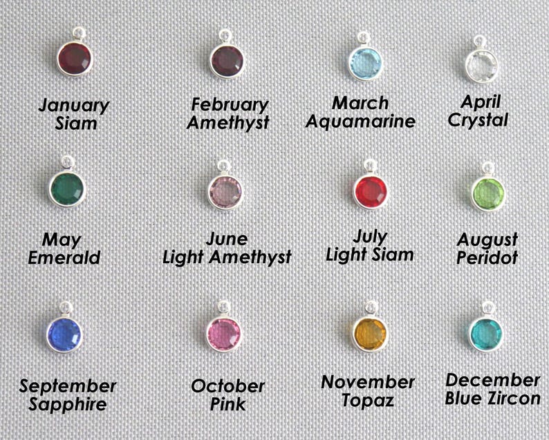 May include: A collection of twelve circular silver birthstone charms arranged in a grid. Each charm features a coloured crystal and is labelled with its corresponding month and stone name, including January Siam, February Amethyst, March Aquamarine, April Crystal, May Emerald, June Light Amethyst, July Light Siam, August Peridot, September Sapphire, October Pink, November Topaz, and December Blue Zircon.