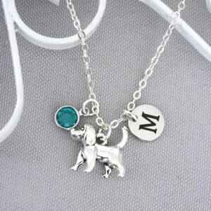 May include: A silver-toned necklace featuring a dog-shaped charm, a circular charm engraved with the letter M, and a small round teal-colored crystal charm. The charms are suspended from a delicate silver-toned metal chain link necklace.