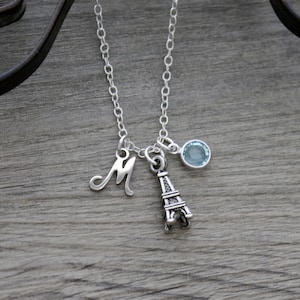 May include: A silver necklace with a charm of the Eiffel Tower, a letter 'M' charm, and a light blue gemstone.