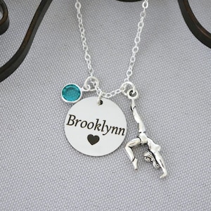 May include: Silver necklace with a personalized disc charm engraved with the name "Brooklynn" and a small heart. The necklace also features a charm of a gymnast performing a handstand.