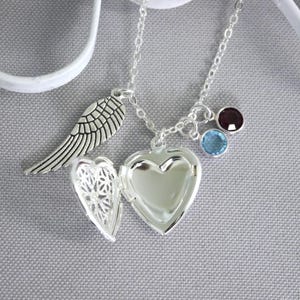 May include: A silver necklace with a heart-shaped locket, a small angel wing charm, and three gemstone charms: one dark purple and two light blue.