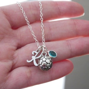 May include: Silver necklace with a soccer ball charm, a letter K charm, and a green gemstone.