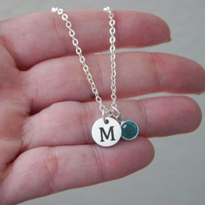 May include: A silver necklace with a delicate chain. It features a round pendant with the letter "M" and a small, teal-colored gemstone charm. The necklace is displayed against a neutral background.