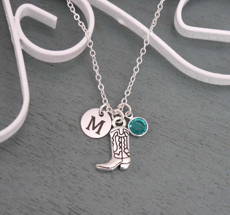 May include: Silver-toned necklace with three charms: a round disc with the letter "M", a cowboy boot, and a teal-colored gemstone. The necklace is displayed against a gray background.