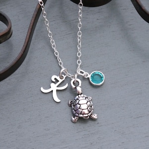 May include: A silver-toned necklace featuring a delicate chain with three charms: a silver letter K initial, a small silver-toned sea turtle pendant with detailed shell markings, and a single round, faceted turquoise-colored crystal bead.