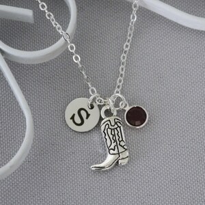 May include: Silver-toned necklace featuring a charm with the letter "S", a cowboy boot, and a small, round, dark red gemstone. The chain is delicate, and the charms hang from it. The background is a light gray.