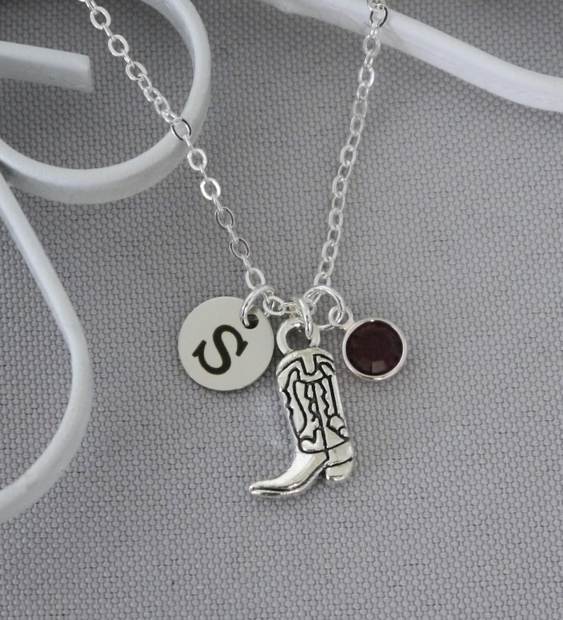 May include: Silver-toned necklace featuring a charm with the letter "S", a cowboy boot, and a small, round, dark red gemstone. The chain is delicate, and the charms hang from it. The background is a light gray.