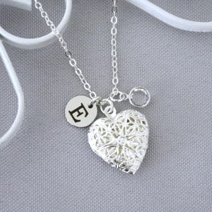 May include: A silver-colored heart-shaped locket necklace with a delicate filigree design. The necklace includes a small round charm with the letter "E" and hangs on a silver chain. The locket is approximately 1 inch in height.