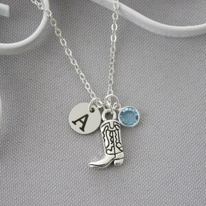 May include: Silver-toned necklace featuring a charm with a cowboy boot design, a round charm with the letter "A", and a light blue gemstone. The necklace is on a silver chain.