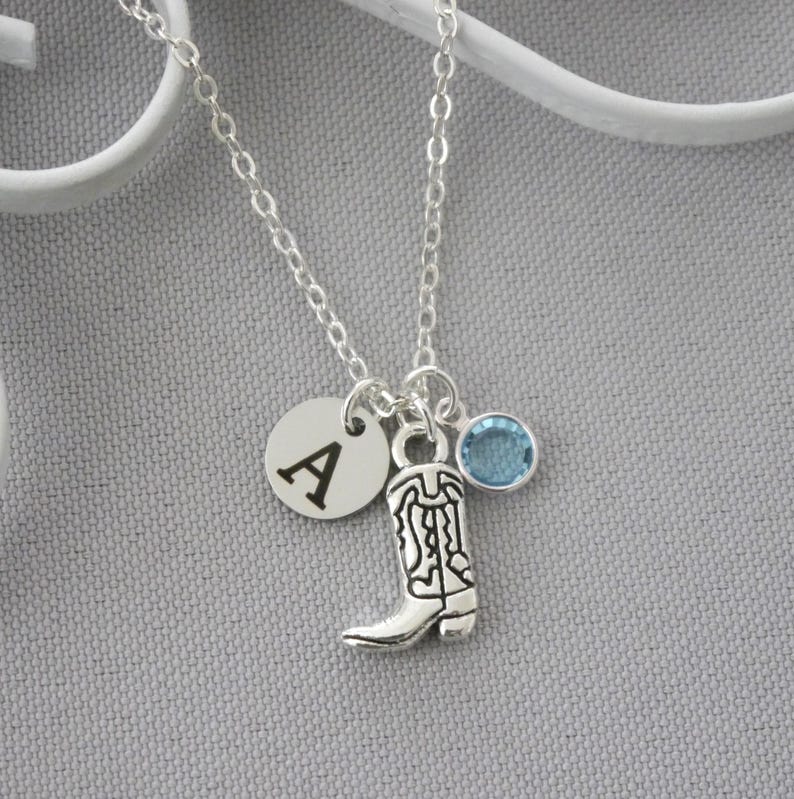 May include: Silver-toned necklace featuring a charm with a cowboy boot design, a round charm with the letter "A", and a light blue gemstone. The necklace is on a silver chain.