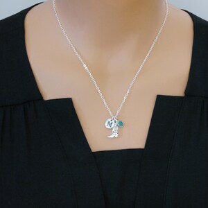 May include: Silver necklace with a small cowboy boot charm, a round charm with the letter "M", and a teal-colored charm. The necklace is worn against a black top.