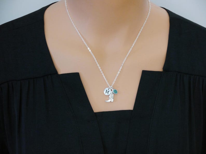 May include: Silver necklace with a small cowboy boot charm, a round charm with the letter "M", and a teal-colored charm. The necklace is worn against a black top.