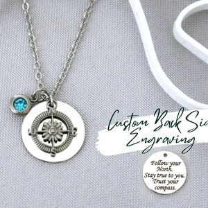May include: A silver-toned necklace featuring a circular pendant with a compass design and a small blue crystal charm. A separate circular silver disc displays the engraved text: Follow your North. Stay true to you. Trust your compass.