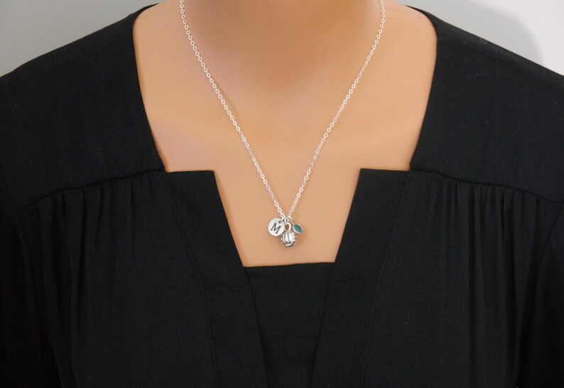 May include: A silver-toned chain necklace featuring a small circular charm engraved with the letter M, a miniature silver-toned volleyball charm, and a small, round, teal-colored accent bead hanging from the center of the delicate metal chain.