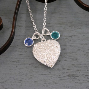 May include: A silver heart-shaped locket necklace with a delicate filigree design. The necklace features two small gemstone accents, one blue and one teal.