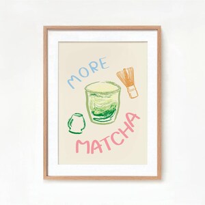 May include: A framed print with a light beige background featuring a green matcha drink in a glass with a green tea whisk and the words "More Matcha" in pink and blue.