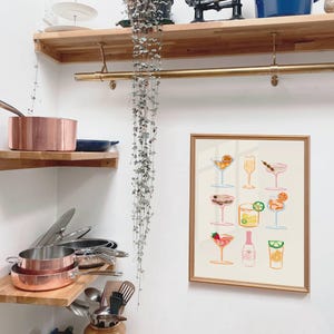 May include: A gold framed print with a white background and a colorful illustration of 12 different cocktails. The print is hanging on a white wall above a set of wooden shelves.