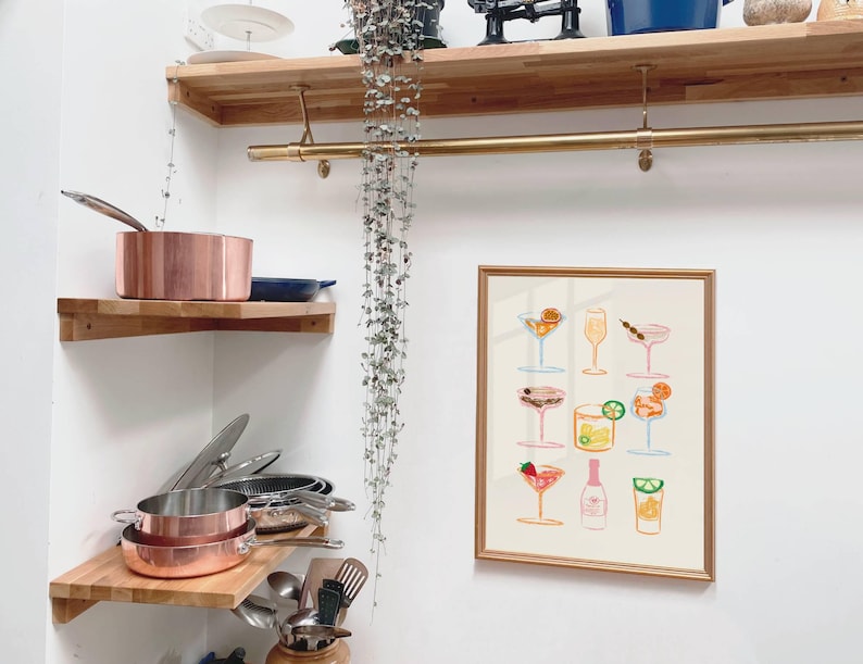 May include: A gold framed print with a white background and a colorful illustration of 12 different cocktails. The print is hanging on a white wall above a set of wooden shelves.
