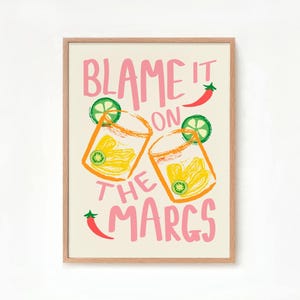 May include: A framed print with the text "Blame it on the Margs" in pink lettering. The print features two illustrations of margarita glasses with lime wedges and a red chili pepper.