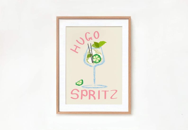Op de afbeelding: Een ingelijste print met een lichtbeige achtergrond met een handgetekend ontwerp van een cocktailglas met een limoenpartje en een munttakje. De tekst "HUGO SPRITZ" is geschreven in roze letters.