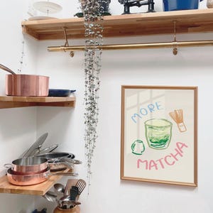 May include: A framed illustration of a green matcha drink with a whisk and the text "More Matcha". The illustration is on a white background with a gold frame. The frame is hanging on a white wall.
