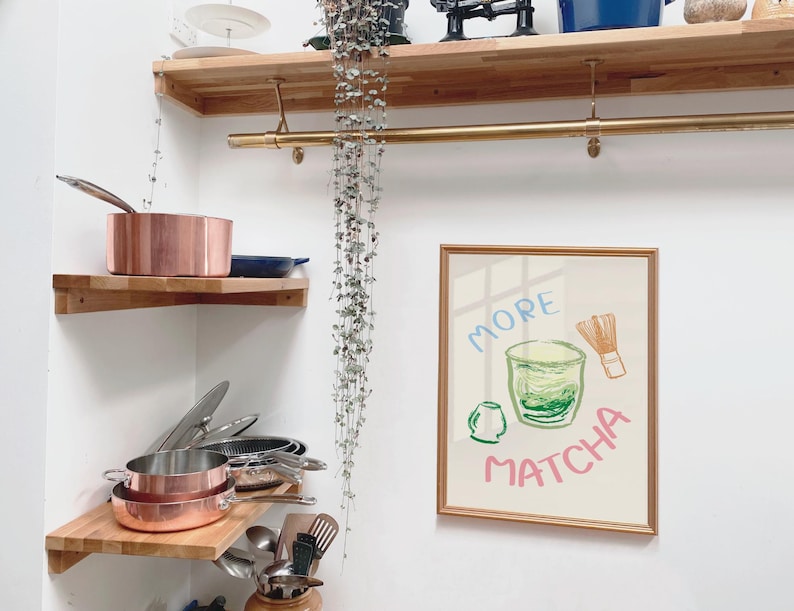 May include: A framed illustration of a green matcha drink with a whisk and the text "More Matcha". The illustration is on a white background with a gold frame. The frame is hanging on a white wall.