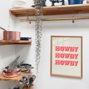 May include: A gold framed print with the text "Howdy Howdy Howdy" in pink letters on a white background. The print is hanging on a white wall in a kitchen with wooden shelves and copper pots.