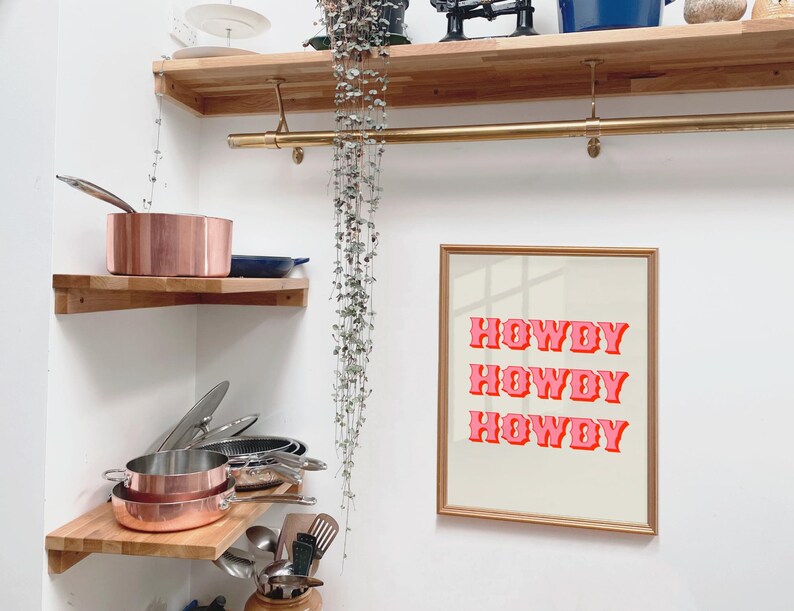 May include: A gold framed print with the text "Howdy Howdy Howdy" in pink letters on a white background. The print is hanging on a white wall in a kitchen with wooden shelves and copper pots.