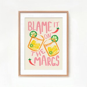 May include: A framed print with a light beige background and pink text that reads "Blame it on the Margs". The print features two illustrations of margarita glasses with lime wedges and a red chili pepper.