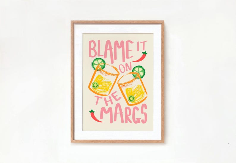 May include: A framed print with a light beige background and pink text that reads "Blame it on the Margs". The print features two illustrations of margarita glasses with lime wedges and a red chili pepper.