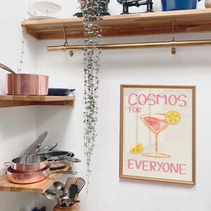 May include: A wooden shelf with a gold metal rod above it. The shelf is filled with copper pots and pans. A framed print with the text "Cosmos for Everyone" and a pink cocktail glass with a yellow lemon slice is hanging on the wall.