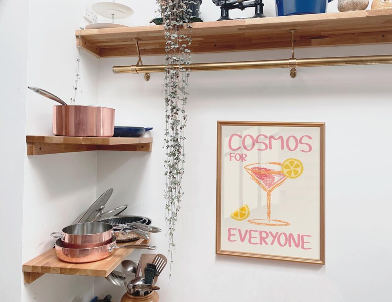 May include: A wooden shelf with a gold metal rod above it. The shelf is filled with copper pots and pans. A framed print with the text "Cosmos for Everyone" and a pink cocktail glass with a yellow lemon slice is hanging on the wall.