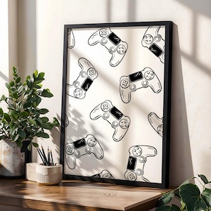 May include: A framed print displaying a pattern of black and white video game controllers. The controllers are repeated on a white background, within a black frame. The print is on a wooden surface, beside a potted plant and pencils.