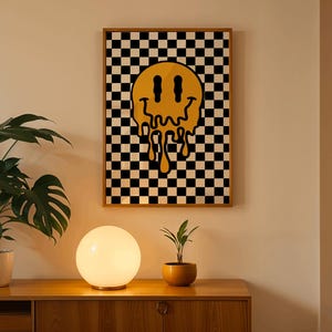 Può includere: Una stampa artistica da parete incorniciata con una faccina sorridente gialla che si scioglie su uno sfondo a scacchi in bianco e nero. L'opera d'arte è esposta sopra un mobile in legno con una lampada a sfera e piante in vaso.