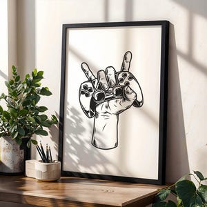 May include: Black framed art print showing a hand making the rock on gesture, holding two video game controllers. The illustration is in black and white. The print is displayed on a wooden surface with plants and pencils.