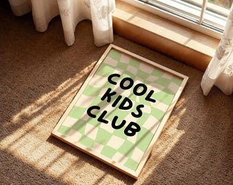 May include: A framed print with the text "COOL KIDS CLUB" in black, set against a light green and cream checkered background. The frame is a light wood color. The print is lying on a brown carpet near a window.