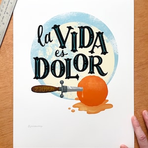 以下が含まれることがあります： 白い背景に青い円と黒い文字で「la VIDA es DOLOR」と書かれています。オレンジにナイフが刺さっており、オレンジジュースが垂れています。