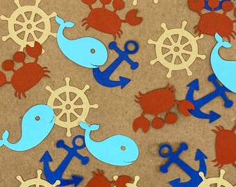 May include: A collection of nautical-themed paper cutouts scattered on a brown surface. The cutouts include blue anchors, light blue whales, tan ship wheels, and rust-colored crabs. The shapes are evenly distributed across the background.