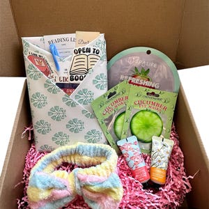 May include: A brown cardboard box filled with a self-care gift set. The box contains a headband, two cucumber eye masks, two hand creams, and a reading list. The reading list is folded into a paper envelope with a green and white floral pattern. The box is filled with pink crinkle paper.