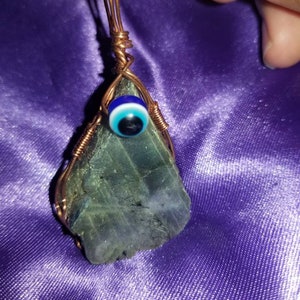 May include: A copper wire wrapped pendant featuring a green labradorite stone with an evil eye charm. The pendant is hanging from a copper wire loop.
