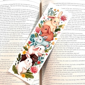 May include: A whimsical bookmark featuring a stack of watercolor bunnies in various colors, surrounded by flowers and greenery. The bookmark is set against an open book with text.
