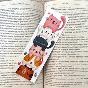 May include: A vertical bookmark featuring a stack of five illustrated cats in pink, white, black, light pink, and brown colors. The cats are arranged vertically with small red hearts and green leaf accents. The bookmark rests on an open book page.
