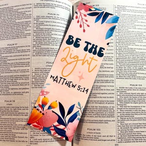 May include: A colorful bookmark with the text "BE THE LIGHT MATTHEW 5:14" is placed inside an open book. The bookmark features floral and leaf designs in pink, blue, and yellow. The book's pages display printed text.